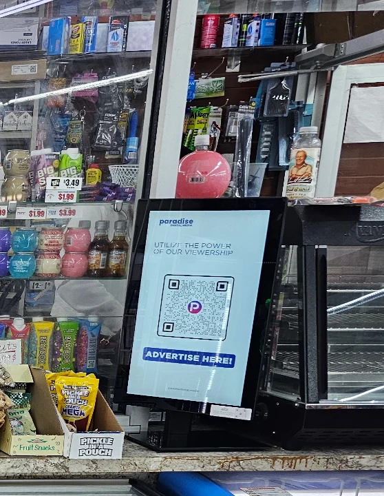 A convenience-store counter: a countertop display stands between the register and a heated case, with the store's shelves behind it.